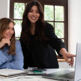 Zwei lächelnde Frauen arbeiten gemeinsam an einem Laptop in einem hellen Büro. Eine sitzt am Schreibtisch, die andere steht daneben und zeigt auf den Bildschirm. Im Hintergrund sind Fenster mit Bäumen zu sehen.