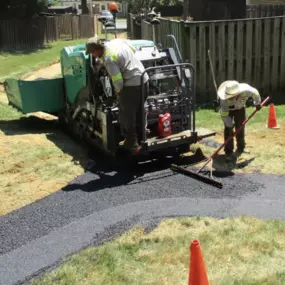 Two workers in safety gear operate paving machinery to lay down fresh asphalt on a grassy area.