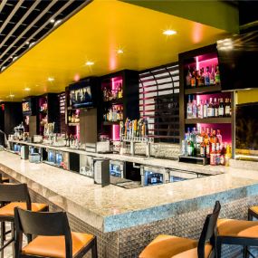 The image shows a bar with a friendly bartender standing behind the counter. The bar has lots of different alcohol bottles neatly arranged on shelves. The place looks inviting with warm lights shining on the various drinks.