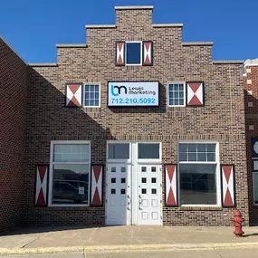 Our Front showcases a brick commercial building with a symmetrical facade, featuring white double doors and large windows on either side. The building has distinctive red and white decorative accents, giving it a charming, vintage appeal. A sign above the entrance prominently displays the logo and contact information for 