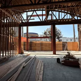 A partially constructed building featuring an open structure with visible wooden framing and steel beams. The bright daylight filters through the incomplete walls, showcasing the scale and framework of the construction. The site is surrounded by temporary wooden barriers, with a view of neighboring buildings and a blue sky in the background.