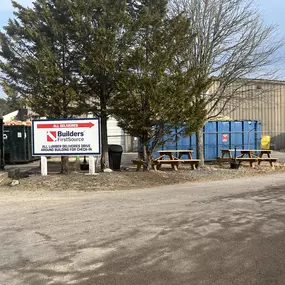 Builders FirstSource sign directing all deliveries, including lumber shipments, to drive around the building for check-in. The outdoor setting includes dumpsters, picnic tables, and warehouse buildings. Located on Welby Road, Massachusetts.