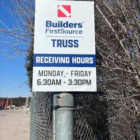 A close-up of a Builders FirstSource sign displaying truss receiving hours, indicating operational times from Monday to Friday, 6:30 AM – 3:30 PM. The sign is mounted on a metal post along a chain-link fence, with a construction yard in the background.