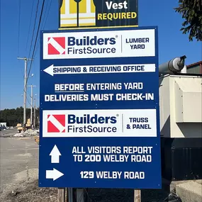 A safety sign at Builders FirstSource requiring all personnel to wear a safety vest beyond this point. The sign provides directions to the lumber yard, truss & panel division, and shipping & receiving office. Located at 129 and 200 Welby Road, Massachusetts.