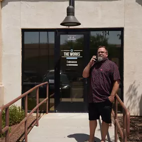 In this image, Mike Rux, a seasoned professional at Ciphers Digital Marketing, is seen outside the Gilbert, Arizona office. He is dressed casually in a patterned short-sleeve shirt and black shorts, standing on a ramp leading to the building entrance marked 