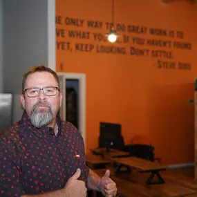 In this image, Mike Rux, a professional from Ciphers Digital Marketing, is seen indoors, giving two thumbs up with a confident and motivated expression. He is wearing glasses and a patterned short-sleeve shirt. The setting features an orange wall with a prominent motivational quote by Steve Jobs: 