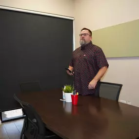 In this image, Mike Rux, a professional at Ciphers Digital Marketing, is seen standing at the head of a conference table during a session on social media marketing at their Gilbert, Arizona office. He is holding a phone and wearing glasses and a patterned short-sleeve shirt. The conference room features a black window shade, a potted plant, and office supplies arranged neatly on the table, creating a professional and organized environment. Mike's attentive and thoughtful demeanor highlights his 