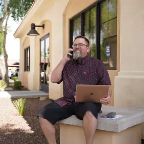 In this image, Mike Rux, a professional from Ciphers Digital Marketing, is captured outside the office in Gilbert, Arizona. He is sitting comfortably on a concrete bench, dressed in a casual patterned short-sleeve shirt and black shorts. With a laptop on his lap, Mike engages in a phone conversation, smiling and exuding a friendly and approachable demeanor. The building behind him features large windows and a light-colored exterior, and the surroundings include a well-maintained sidewalk and lan