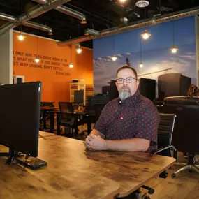 In this image, Mike Rux, an SEO professional at Ciphers Digital Marketing, is seated at a wooden desk in their Gilbert, Arizona office. He is wearing glasses and a patterned short-sleeve shirt, and his serious expression indicates focus and professionalism. The office environment includes computer monitors on wooden desks, an orange wall featuring a motivational quote by Steve Jobs, and a large mural of a scenic landscape. Other team members are seen working in the background, creating a collabo