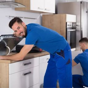 Zwei Männer in blauen Arbeitsuniformen installieren Küchengeräte in einer modernen Küche. Der Mann im Vordergrund schaut lächelnd in die Kamera, während er an einem Küchengerät arbeitet. Die Szene unterstreicht die fachkundige Demontage- und Montagedienste, die WIR ENTSORGEN anbietet.