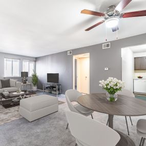 A dining room with a ceiling fan leads to a living room with two windows near a hallway and a kitchen.