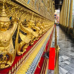 A stunning close-up of intricate golden Garuda statues, each carrying a serpent, lining the exterior wall of a temple in Thailand's Grand Palace. The craftsmanship showcases detailed ornamentation, rich gold tones, and a sense of cultural significance. The polished marble floor and colorful, mirrored mosaics further highlight the elegance of traditional Thai architecture. This image captures the grandeur and heritage of Southeast Asia, making it a prime destination for immersive luxury travel ex