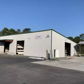 A large metal warehouse building with two open bays is visible. The building has beige siding and a green roof, with stacks of building materials like wood and other supplies stored inside. A sign on the side of the building reads 