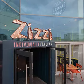 Exterior view of Zizzi Liverpool One, showing modern glass front and outdoor seating area surrounded by planted greenery.
