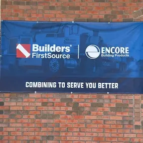 A large banner displayed on a brick wall with the logos of Builders FirstSource and Encore Building Products. The banner has a blue background with an image of a truck in the background and white text that reads, 