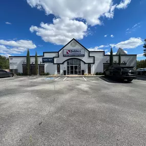 he exterior of a Builders FirstSource showroom with a white facade and dark trim. The entrance features glass double doors beneath a large sign displaying the Builders FirstSource logo and the words 