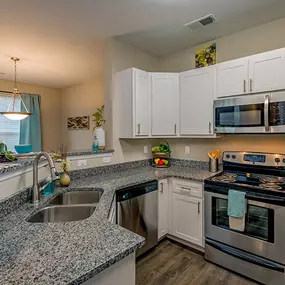 A kitchen with granite countertops, stainless steel appliances, hardwood-style flooring, a double basin sink, and white cabinets near a dining room.