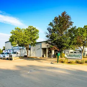 The image depicts the exterior of Greenwood Cleaning Systems, located in Davenport, Iowa. The building is a light industrial facility with a white metal façade, surrounded by neatly landscaped greenery, including several trees and trimmed bushes. The Greenwood Cleaning Systems sign is prominently displayed in the foreground on a green post, adding to the professional appearance. The parking area is clean and spacious, with company-branded trucks parked near the entrance, showcasing their operati