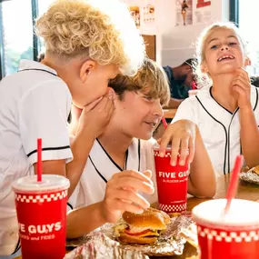 Children chat and laugh while enjoying their Five Guys meals at a dining room table in a Five Guys restaurant.