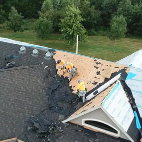 Close-up aerial view of a roofing crew working on a steep section of a house’s roof. Workers in bright yellow shirts are removing old shingles and preparing the roof for new installation using CertainTeed underlayment. The surrounding area includes a backdrop of green lawns and trees, emphasizing the rural location.