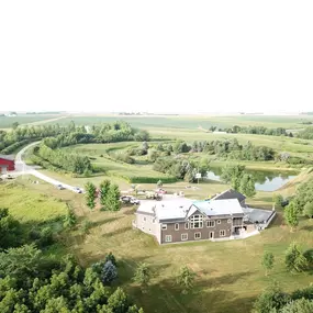 Aerial view of a countryside property featuring a modern two-story home with ongoing roofing work. The home is surrounded by rolling farmland, a scenic pond, and lush greenery. A bright red barn stands prominently to the left, connected by a winding driveway that adds a charming rural touch.