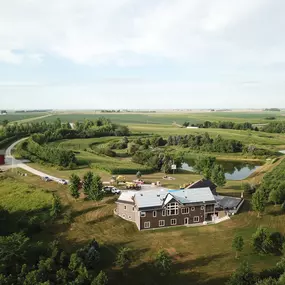 A breathtaking aerial view of a modern two-story home nestled in a picturesque rural setting. The property features expansive green lawns, a tranquil pond, and a bright red barn visible near a winding driveway. The roof of the home is undergoing renovations, with workers and construction equipment onsite, blending the process of improvement with the natural beauty of the surrounding farmland.