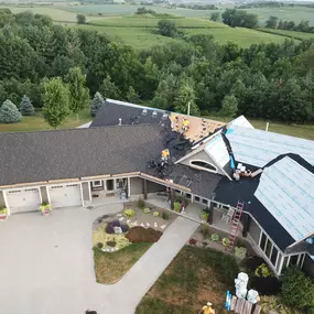 A detailed aerial view of roofing work on a modern single-story home with a large garage and landscaped front yard. Workers are seen actively removing old shingles and installing new materials, including CertainTeed underlayment. The property is surrounded by lush greenery and rolling farmland, offering a serene rural backdrop.