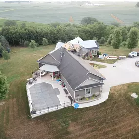 Aerial view of a modern home with an expansive backyard featuring a fenced-in pool and a large patio. Roofing work is in progress, with visible construction materials and workers actively replacing shingles. The surrounding property includes neatly mowed lawns, trees, and a wide driveway leading to the house and garage. The rural farmland beyond adds a picturesque backdrop.