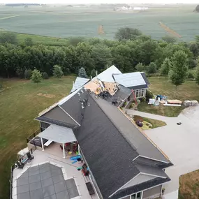 Aerial view of a modern home under roof replacement, with workers actively repairing the shingles on multiple sections of the roof. The property features a spacious patio, a fenced pool, and a large driveway. The well-manicured lawn and surrounding trees blend seamlessly into the vast farmland beyond.