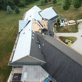 Overhead view of a home’s roof replacement in progress, showing workers actively removing old shingles and applying CertainTeed underlayment. The detailed process is visible on different sections of the roof. Nearby, construction materials, tools, and vehicles are arranged in an organized manner on the lawn and driveway.