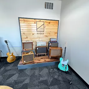 This photo features a rustic display of Sound Salvage amplifiers, arranged against a wooden backdrop with the company’s engraved logo. A mix of amplifiers and a vibrant teal electric guitar are displayed, showcasing unique craftsmanship and design.