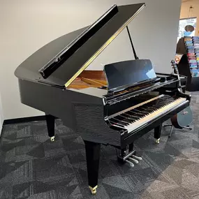 This image features a polished black grand piano displayed in the Brock Family Music showroom, surrounded by guitars and other instruments. The piano's open lid and sleek design make it a focal point in the space.