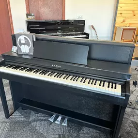 This image shows a sleek Kawai digital piano displayed in the Brock Family Music showroom. The piano features a matte black finish, and its compact design includes headphones and a music stand for practical use. In the background, more pianos and amplifiers can be seen, blending traditional and digital instruments.