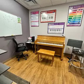 This image shows another music lesson room at Brock Family Music, featuring a wooden upright piano, a stool, a chair, and a large whiteboard. The walls are adorned with colorful educational posters covering musical notes, chords, and keys.