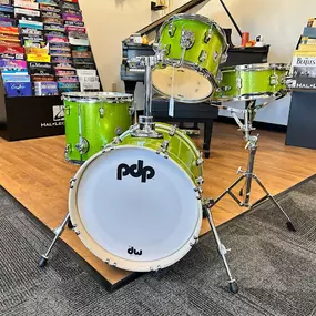 This image showcases a bright green PDP drum set on display in the Brock Family Music showroom. The set includes a bass drum, toms, and a snare, all arranged on a raised wooden platform near shelves of sheet music and books.