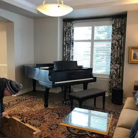 This image showcases a Brock Family Music team member carefully preparing to move a grand piano in a beautifully decorated living room. The piano sits on an ornate rug, with sunlight streaming through large windows in the background.