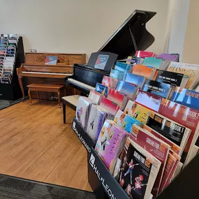 This image showcases a collection of sheet music racks alongside a wooden upright piano and a polished black grand piano. The sheet music includes books for beginners, classical pieces, and popular songs.