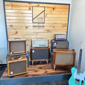 This photo features a rustic display of vintage-style amplifiers from Sound Salvage, set against a wooden backdrop with the company’s logo engraved. The amplifiers are displayed in various sizes and finishes, creating a visually appealing collection. A vibrant teal electric guitar is also showcased alongside the amplifiers, adding a modern pop of color.