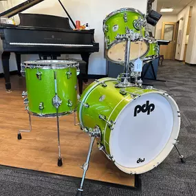 This photo showcases a vibrant green PDP drum set on display in the Brock Family Music showroom. Positioned on a wooden platform with a grand piano in the background, the set includes a bass drum, toms, and a snare drum.