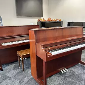 This image features a pair of polished wood Kawai upright pianos, displayed prominently in the Brock Family Music showroom. The warm wood finish adds a touch of sophistication, with matching benches completing the elegant setup. Other upright pianos in polished black can be seen in the background, offering variety for customers to explore.