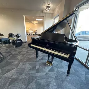 This image captures a polished black grand piano displayed in a spacious, light-filled corner of the Brock Family Music showroom. The piano’s elegant design and open lid emphasize its premium craftsmanship, while the surrounding instruments add to the inviting atmosphere.