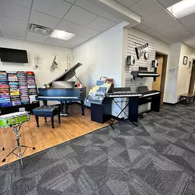 This image provides an overview of the Brock Family Music showroom, featuring a variety of instruments, including a bright green drum set, a grand piano, digital pianos, and shelves of sheet music and learning materials.