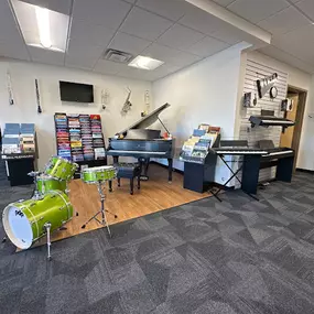 This image captures a section of the Brock Family Music showroom, featuring a green PDP drum set, a grand piano, digital keyboards, and shelves stocked with sheet music. Instruments are displayed on wooden platforms, emphasizing both variety and organization.