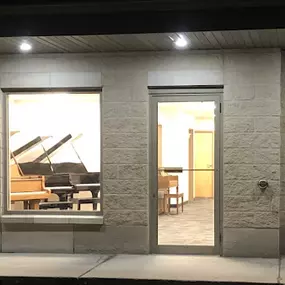 This image shows the exterior of Brock Family Music at night. The brightly lit windows reveal grand pianos on display, inviting passersby to explore the showroom. The warm lighting adds a sense of professionalism and accessibility.