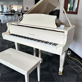 This image showcases a pristine white Kawai grand piano paired with a matching bench, displayed in a spacious area of the showroom. The piano’s elegant design and glossy finish create a striking visual impression.