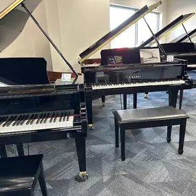 This image showcases a showroom of polished black Kawai grand pianos, arranged in a bright, modern space with large windows that let in natural light. Each piano is displayed with a matching bench, emphasizing their elegance and craftsmanship.