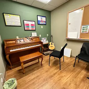 This photo captures a cozy music lesson room at Brock Family Music, featuring a classic upright piano with a wooden finish. The room is equipped with additional seating, a guitar, and colorful teaching posters on the walls, creating an inviting and educational environment.