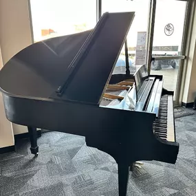 This image highlights a polished black grand piano on display in the Brock Family Music showroom. Positioned near large windows, the piano is bathed in natural light, emphasizing its elegant design and craftsmanship.