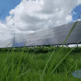 A ground-mounted solar panel array installed in a lush, grassy field under a partly cloudy sky. The solar panels are angled optimally to capture sunlight, supported by a durable metal framework. Tall green grass in the foreground creates a natural, eco-friendly setting that complements the renewable energy technology.