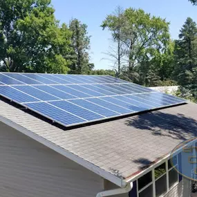 A rooftop solar panel system installed on the sloped roof of a residential home. The solar array is neatly aligned and optimized for maximum sunlight exposure. The house is surrounded by mature trees and a lush green environment, blending the modern solar technology with a natural setting. The bright sunlight highlights the clean and efficient design of the panels.